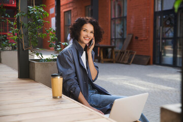 remote workers. Working from Anywhere.  middle aged woman    is working on a laptopon and talking on a mobile phone a bench on the street in a cafe. startup