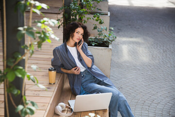  remote workers. Working from Anywhere.  middle aged woman    is working on a laptopon and talking on a mobile phone a bench on the street in a cafe. startup