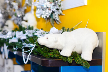 Plush white polar bear toy lounges playfully on shelf, among fresh green Christmas pine branches. Sparkling silver tinsel, ornaments and lush garlands adorn yellow wall behind, evoking holiday cheer