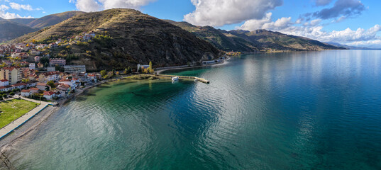 Aerial view of Pogradec, Albania, featuring a calm bay on Lake Ohrid, a small pier, turquoise water, and surrounding hills and mountains.