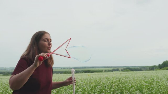 caucasian woman creating giant soap bubble over meadow with red wand, expansive landscape and distant tree line under soft cloudy sky, iridescent orb floats slowly, meditative outdoor art and calm