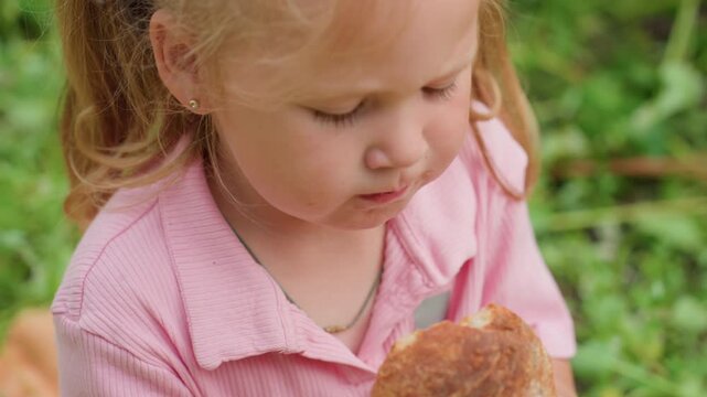 white toddler girl grimacing after bite, rubbing forehead and wiping crumbs from cheek, pausing in thought then offering hesitant smile, contemplative moment under trees during outdoor snack, candid