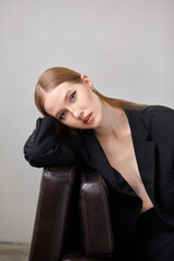 A woman in a black jacket and black trousers is sitting on a brown sofa. She has long brown hair and trendy makeup.