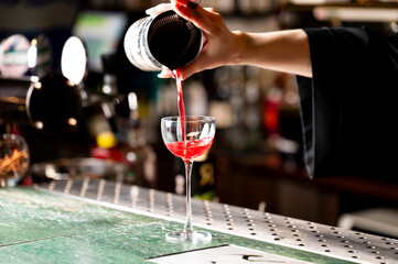 A close-up, eye-level shot of a bartender pouring a vibrant red cocktail from a stainless steel shaker into an elegant coupe glass at a bar with a blurred background