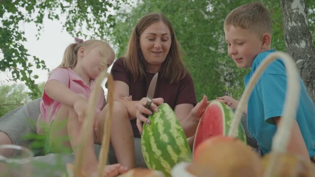 white family picnic sharing watermelon slices outdoors in birch grove mother hands juicy pieces to daughter and son, baskets of fruit in foreground, giggles and sticky hands, sundappled leaves,