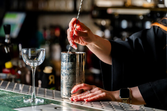 Close-up of a female bartender's hands mixing a cocktail in a silver shaker with a bar spoon at a dimly lit bar counter. The focus is on the drink preparation process - Powered by Adobe