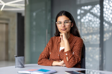 Young south asian businesswoman with glasses sitting at her office desk, looking directly at the...