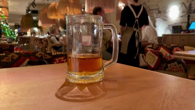 Beer mug on wooden table, amber liquid with reflection, warm cozy restaurant interior, patterned cushions, low pendant lights, server moving in background, shallow depth of field,