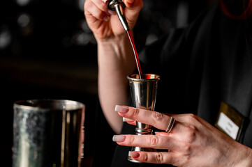 Close-up of a bartender's hands pouring a vibrant red liquid from a bottle into a stainless steel jigger, preparing a cocktail in a dimly lit bar setting