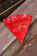 Red hand fan with floral and golden designs placed on wooden surface.