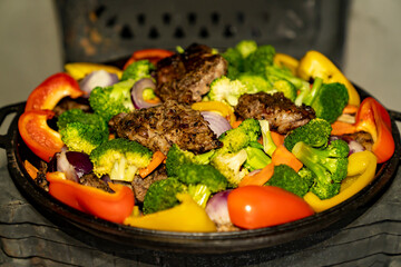 Grilled meat and mixed vegetables cooking in cast iron pan on wood burning stove. Indoor food preparation photography. Home cooking and rustic kitchen concept. Design for poster, banner, wallpaper, pr