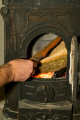 Hand placing firewood inside cast iron wood burning stove with open door and visible flame. Indoor heating process photography. Home heating and solid fuel stove concept. Design for poster, banner, wa