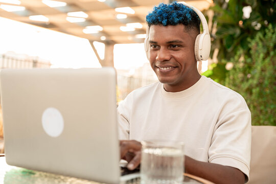 Young latin man with blue hair focused on his laptop, wearing headphones, and smiling while working remotely at an outdoor coffee shop, enjoying flexible work and digital opportunities - Powered by Adobe
