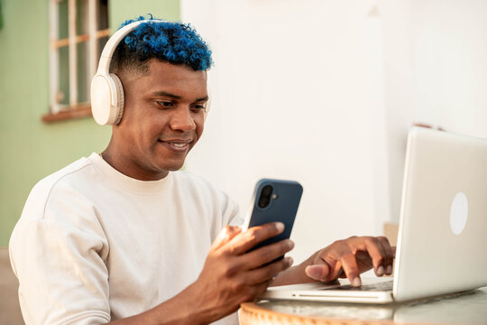 Young latin man with blue hair and headphones enjoying remote work, multitasking on a laptop and smartphone while smiling confidently at an outdoor cafe - Powered by Adobe
