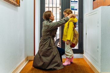 Mother adjusting her daughter's yellow raincoat, preparing kid for school in hallway before leaving home, showing family love, morning routine, and parental care © Dani D.G