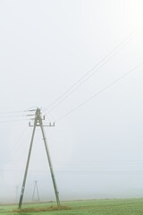 Old concrete utility pole standing in a green agricultural field during heavy fog. Minimalist rural landscape with overhead electric power lines disappearing into the mist. Moody, grey atmosphere