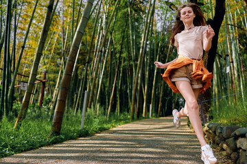 Fototapeta premium Woman jumping on a gravel path in a bamboo forest, casual outfit with sneakers, joyful happiness and motion captured during a summer walk on a green nature trail with sunlight.