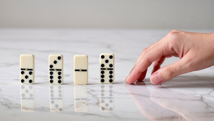 A concise and meaningful composition featuring four white domino tiles standing vertically in a row as a human hand reaches out to trigger the "domino effect." Set on a light marble tabletop with real