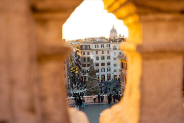 Festive view of Rome near the Spanish Steps seen through historic stone architecture, with a decorated Christmas tree and lively city square below. Winter holiday atmosphere, tourism, city break