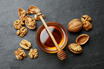 Delicious honey in glass bowl, dipper and walnuts on dark textured table, flat lay