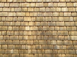 A wall of overlapping wooden shingles with natural grain and warm golden brown tones forms a repeating geometric pattern with rustic texture and weathered details