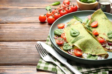 Tasty spinach crepes with tomatoes, onion, sour cream and leaves on wooden table, closeup. Space for text