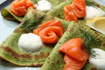 Tasty spinach crepes with salmon, sour cream, dill and lemon on plate, closeup