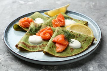 Tasty spinach crepes with salmon, sour cream, dill and lemon on grey table, closeup
