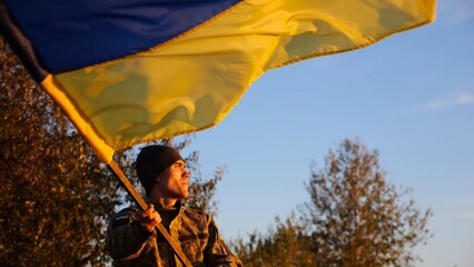 Male military in uniform waving flag of Ukraine at countryside. Young soldier of ukrainian army lifting blue-yellow banner as symbol of victory against russian aggression. Invasion resistance concept
