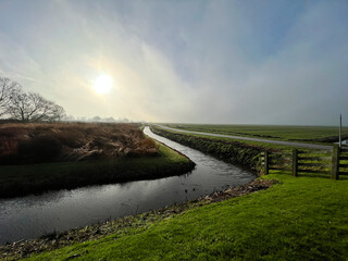 Misty Dutch polder landscape with a calm canal reflecting the bright morning sun, bordered by green grass and a wooden fence.