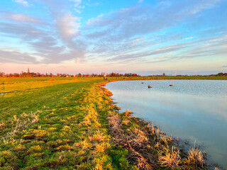 Golden hour Dutch polder landscape with a calm lake, green dike, distant village skyline, and wind turbine.