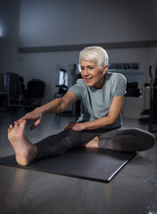 A senior woman stretches her leg on a mat in a gym