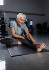 A senior woman stretches her leg on a mat in a gym