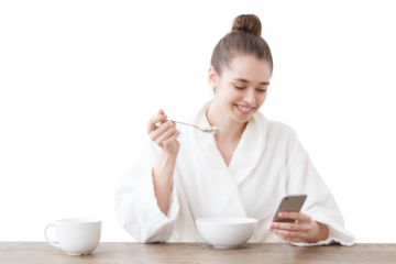 Smiling young woman in a bathrobe checking her mobile phone with calorie tracker app while enjoying a healthy breakfast