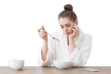 Young woman with no appetite, tired, bad mood and sleepy, sitting in the white loft kitchen, having unsavory unpalatable unappetizing breakfast
