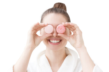 Playful woman in a bathrobe smiling while holding two pink macaron cookies over her eyes
