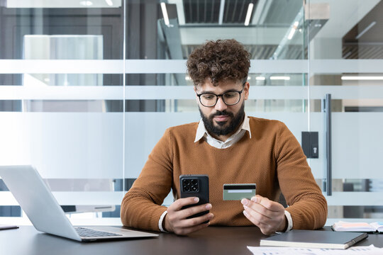 Bearded man holding a smartphone and a credit card. Processing an online payment or making a purchase while sitting at a desk in a modern office setup. Representing e-commerce and financial technology - Powered by Adobe