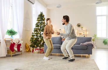 Fototapeta premium Christmas couple dancing disco fun dance at home party near festive Xmas tree with ornaments. Young happy man and woman share joy and laughter beside gifts, stockings and decor in living room