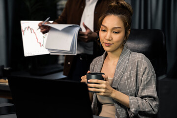 Businesswoman working on laptop with stock market to invest while drinking coffee at modern office while coworker checking paperwork business plan database report meeting at over late night. Infobahn.