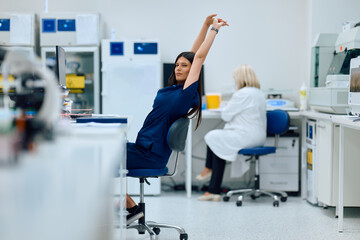 Lab technicians working in a modern laboratory preparing samples while one stretches during a busy workday