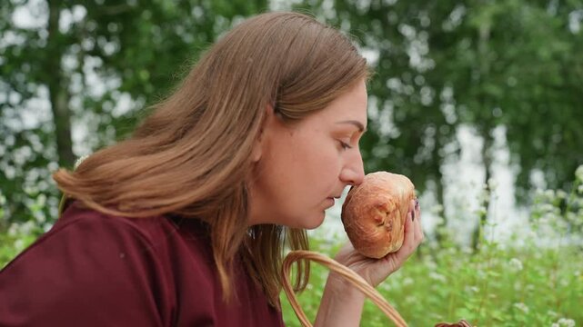 white woman holding loaf near birch trees, smelling fresh bread from woven basket, contemplative expression, wildflower meadow backdrop, maroon shirt, peaceful rural portrait and slow intimate