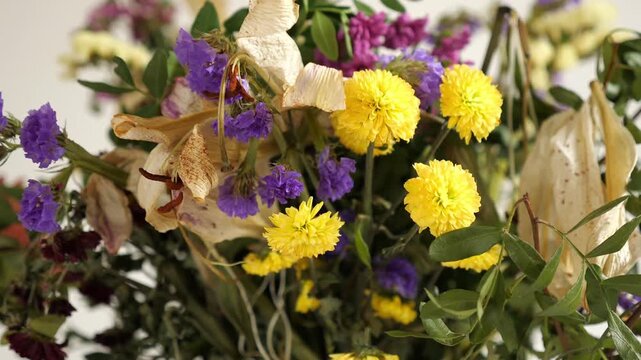 A close-up horizontal indoor shot of a dried flower bouquet with purple Statice (sea lavender), yellow globe amaranth, partially desiccated white lily, and green foliage against a plain, light backgro