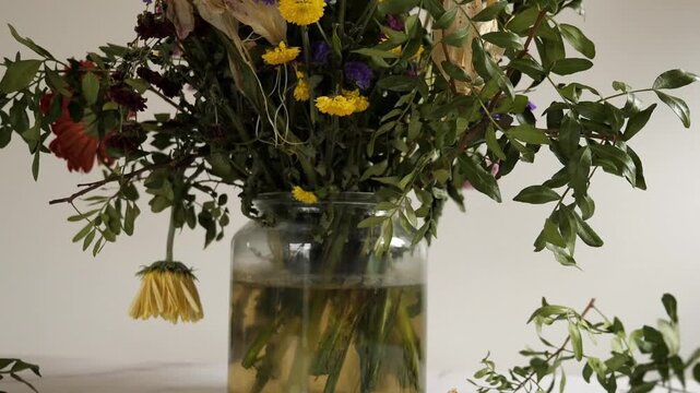 A close-up horizontal indoor shot of a dried flower bouquet with purple Statice
(sea lavender), yellow globe amaranth, partially desiccated white lily, and green
foliage against a plain, light backgro