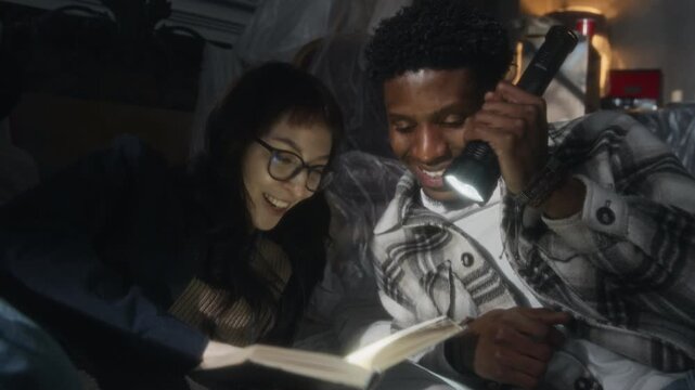 Close up shot of young man and woman reading book together by flashlight talking and laughing while lying on floor of abandoned house together