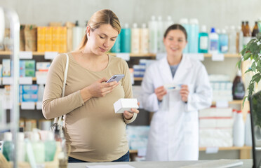 Fototapeta premium Pregnant woman scanning qr code for box of pills with woman pharmacist in pharmacy