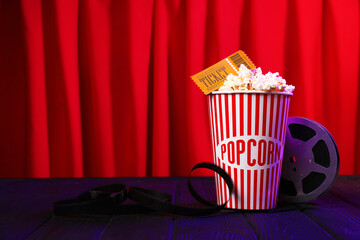 Tasty popcorn in paper cup, movie ticket and film reel on purple wooden table against red curtains, space for text