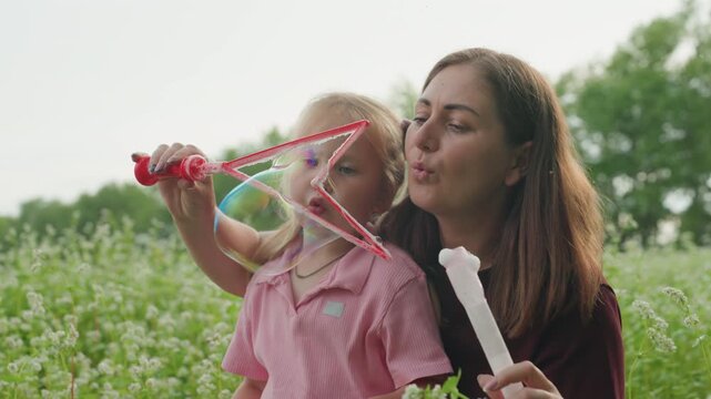 caucasian mother and daughter blowing bubbles in wildflower meadow, intimate teaching moment with pink dress, red bubble wand, soft afternoon light, close embrace and quiet laughter
