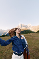 Naklejka premium Woman smiling in denim shirt on mountain meadow outdoors, enjoying nature and breeze. Portrait of happy traveler with jacket and open alpine landscape in soft sunlight.