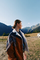 Naklejka premium Woman in profile wearing a patterned coat stands in countryside landscape with mountains in the background and a parked car, autumn travel scene with warm natural light and calm mood.