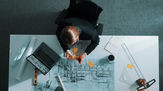 Aerial view of architect drawing while working at laptop. Top down view of caucasian engineer designing blueprint and drafting house model at table with equipment and safety helmet. Alimentation.